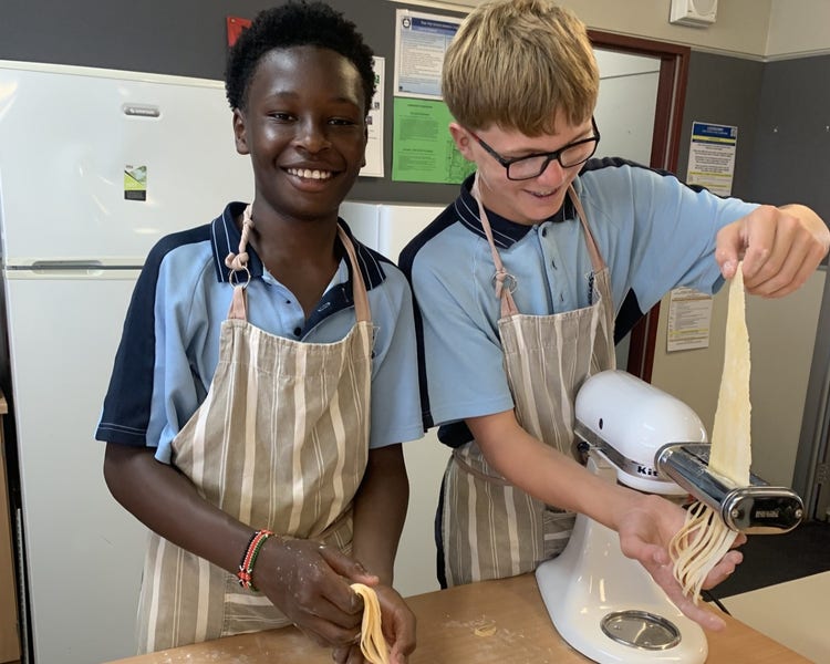 Image of male students making pasta. One student is feeding the pasta through a pasta machine whilst the othe student holds the finished pasta in his hands.