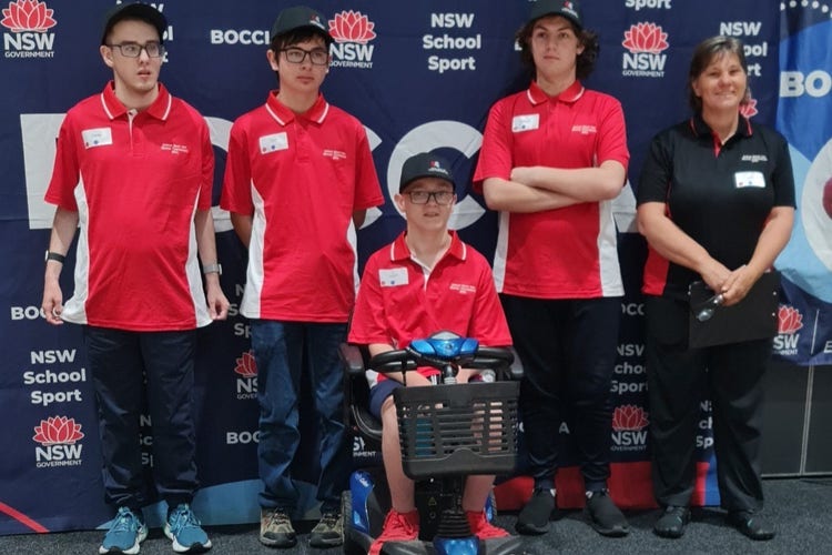 Image of students, one of which is in a mobility scooter and teacher standing in front of the NSW School Sport Boccia signage.