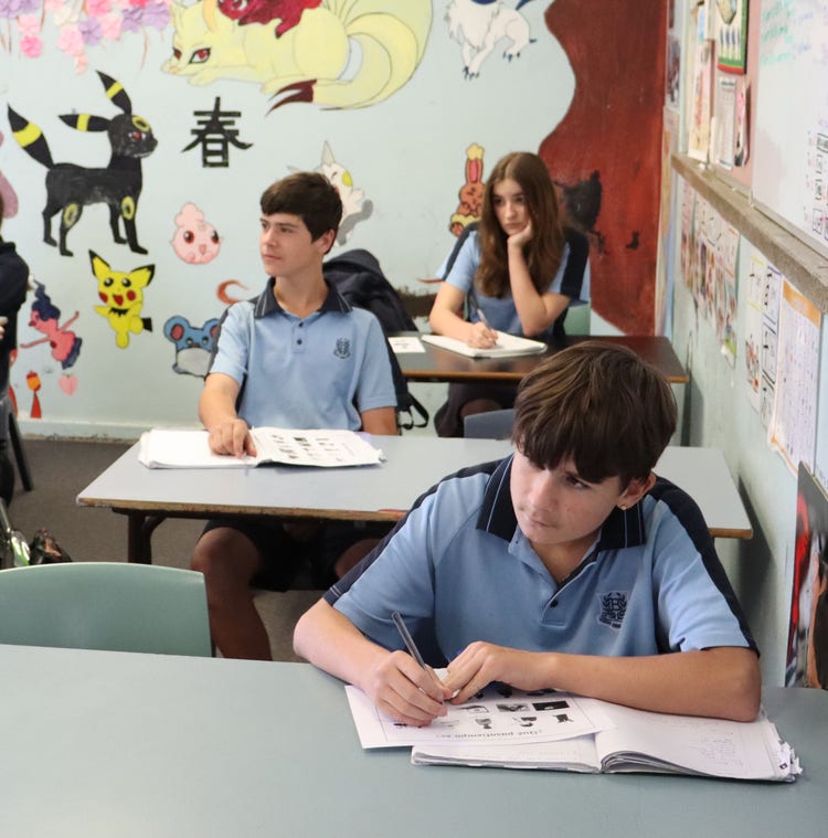 Image of students seated at school desks, writing in open exercise books on the desks. books